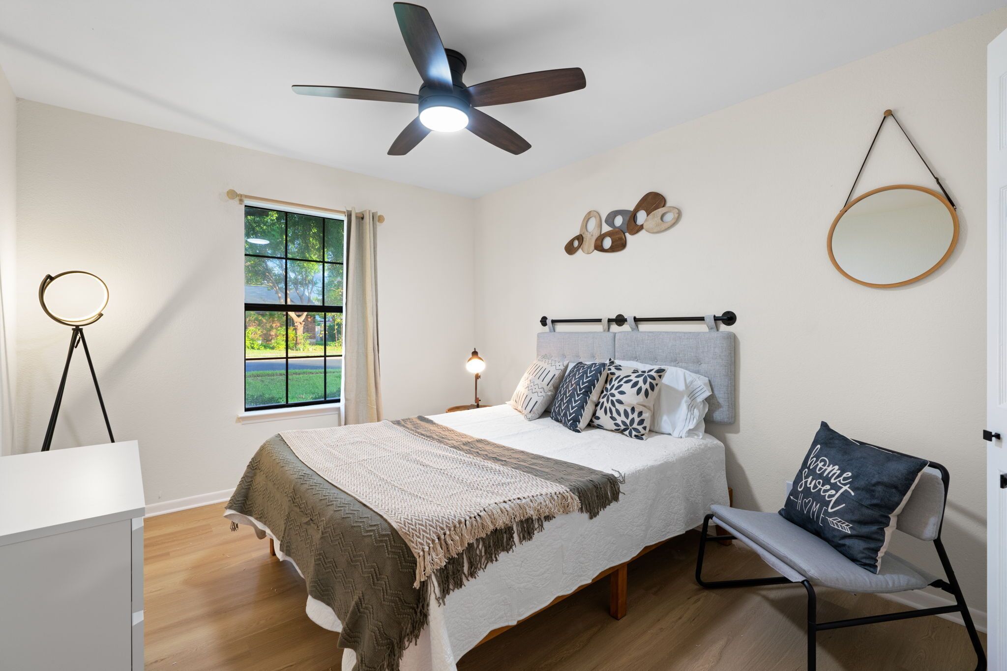 Une chambre à coucher comprenant un lit, une table de chevet, une lampe, une chaise, une fenêtre et un ventilateur de plafond.