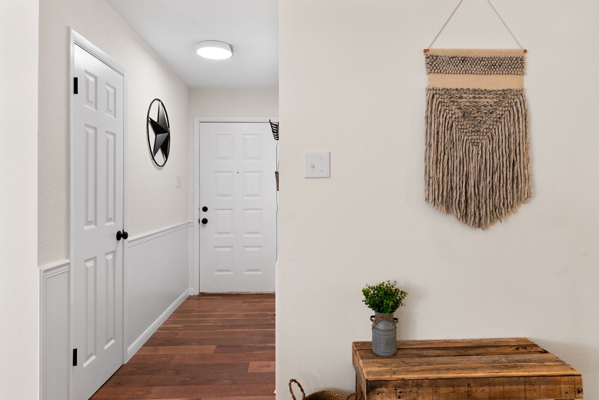 A hallway with white walls, wooden flooring, a white door, a round wall decoration, a macrame wall hanging, and a wooden table with a plant.
