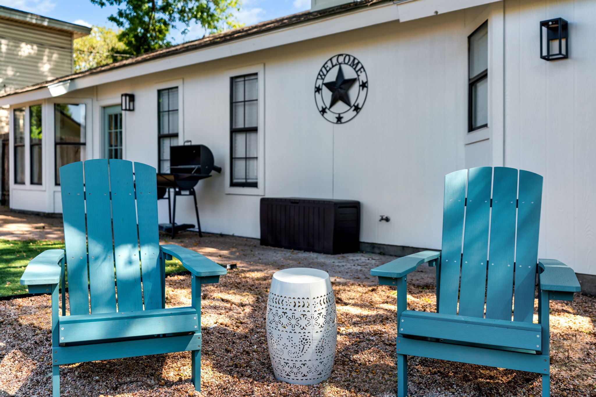Deux chaises Adirondack de couleur bleu sarcelle, un pot de fleurs décoratif blanc et une enseigne « BIENVENUE » ornée d’une étoile, le tout sur la façade extérieure d’un bâtiment blanc.