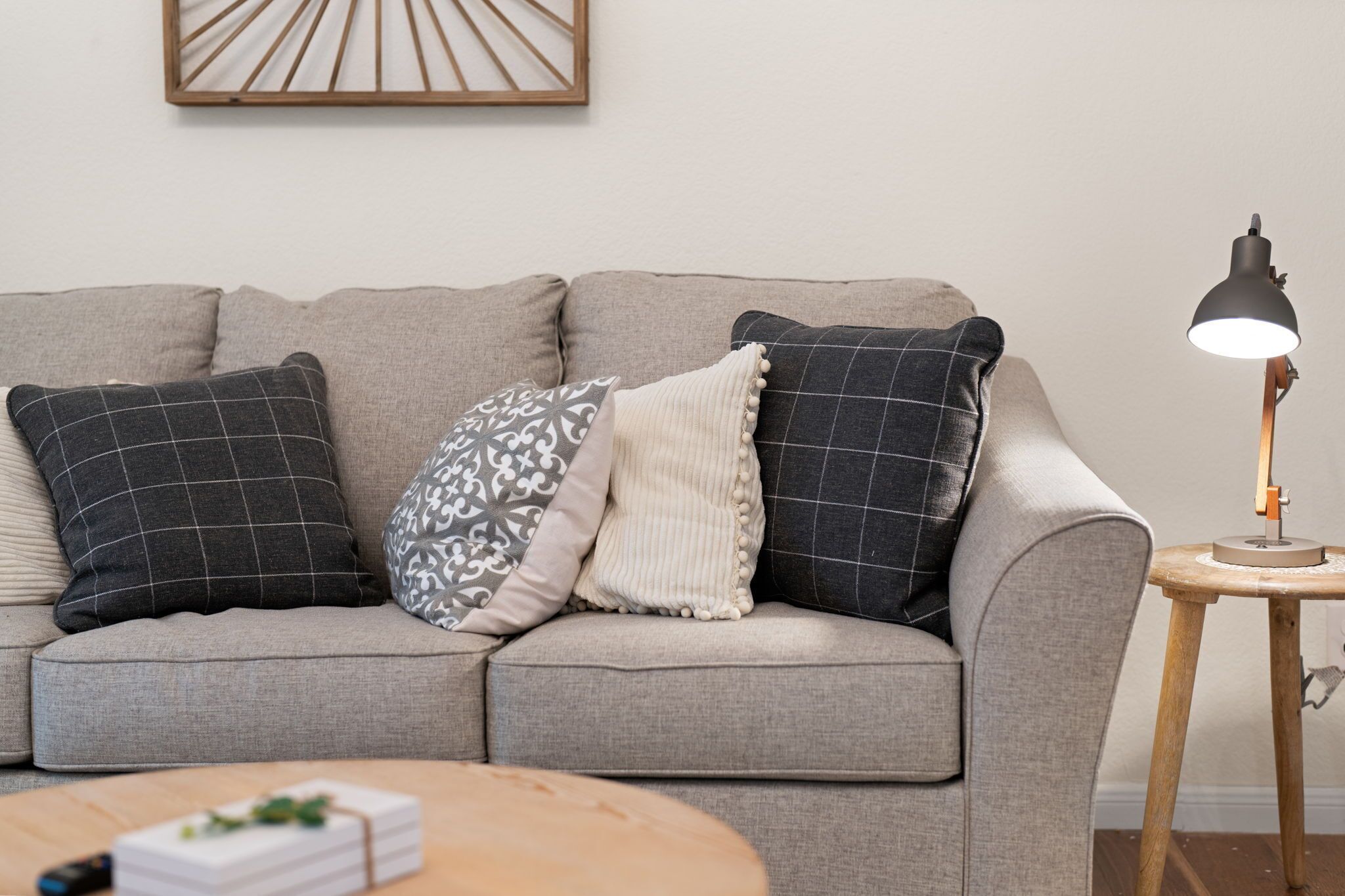 A grey sofa with patterned cushions, a small wooden side table with a lamp, and a framed artwork on the wall.