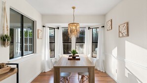 A dining area with a wooden table, white chairs, a chandelier, and large windows with white curtains. - Charming & Bright W Lounging Pool (Round Rock)