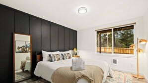 A bedroom with a large bed, a bedside table with a lamp, a window with a view of trees, and a mirror on the wall.