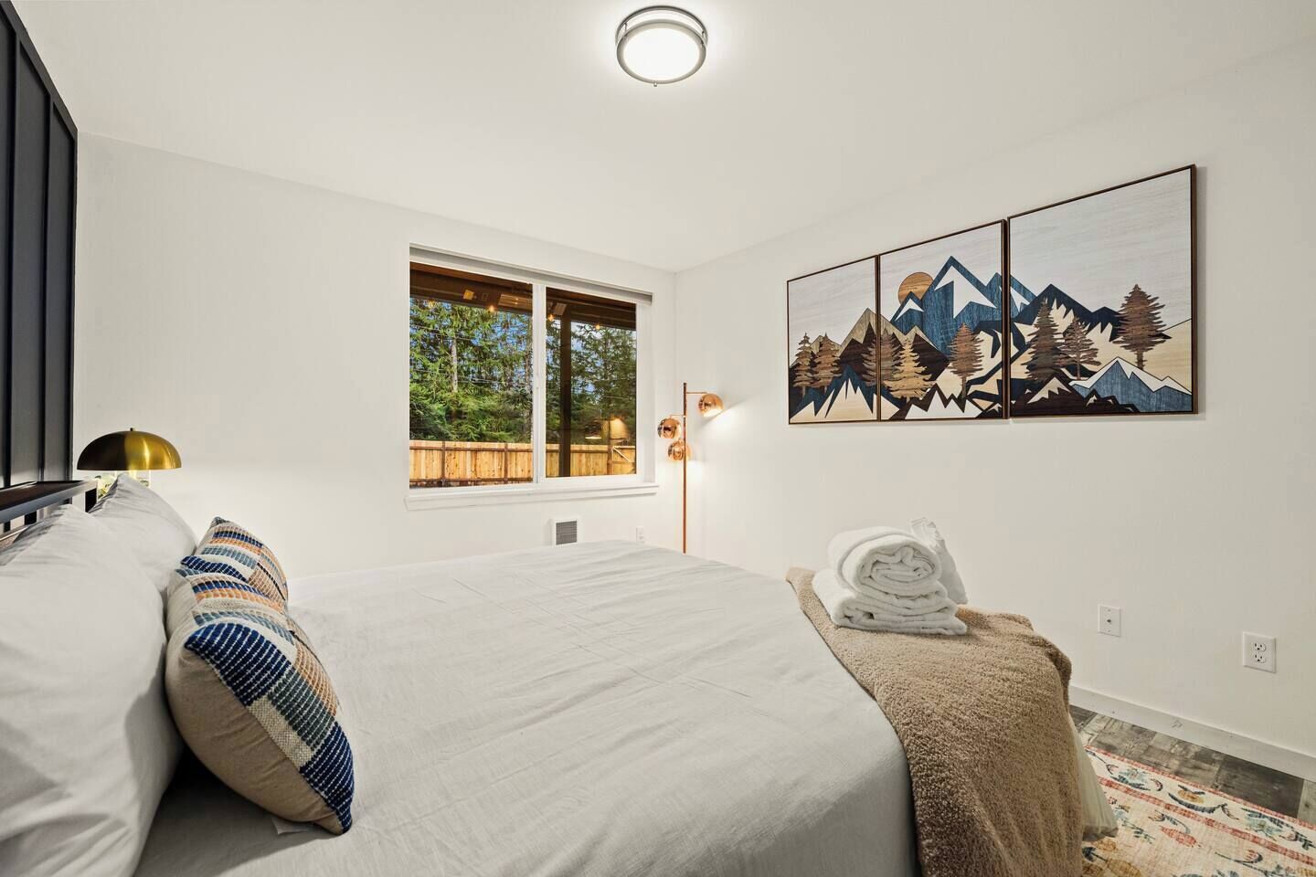 Un dormitorio moderno con una cama grande, una ventana con vistas a árboles, un cuadro de montañas y una lámpara de noche.