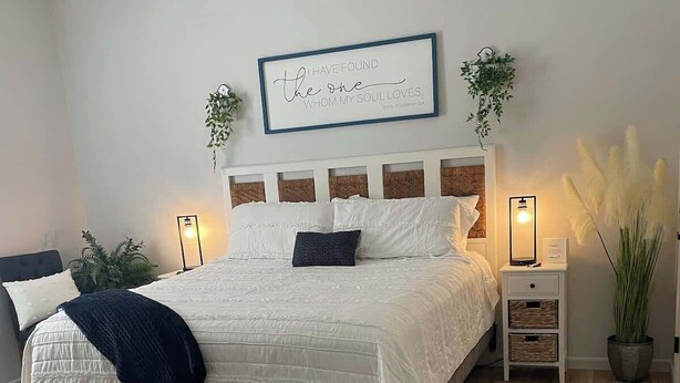 Un dormitorio con una cama grande, mesitas de noche, una lámpara fijada a la pared y una cita enmarcada.