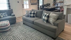 Una sala de estar con un sofá gris, alfombra estampada y un comedor con mesa y sillas.