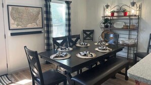 Un comedor con una mesa de madera oscura dispuesta para seis personas, sillas negras, un mapa en la pared y una estantería con plantas en macetas y un farol.