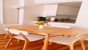 A modern dining area with a wooden table and white chairs, a vase of sunflowers, and a view of the kitchen through an open archway. - Waikiki ocean view studio, easy walk to beach, shopping, restaurants (Honolulu)