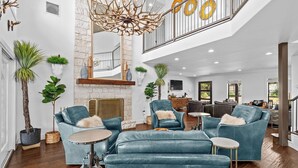 A spacious living area with a stone fireplace, leather sofas, and a high ceiling with a unique chandelier. - Summer Riverfront | 5 Acres | Hot Tub | Sleeps 17 (Wimberley)