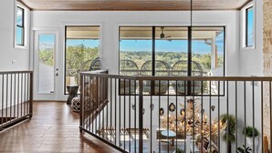 A spacious interior with a wooden ceiling, large windows offering a view of a mountainous landscape, and a chandelier with antler decorations. - Summer Riverfront | 5 Acres | Hot Tub | Sleeps 17 (Wimberley)