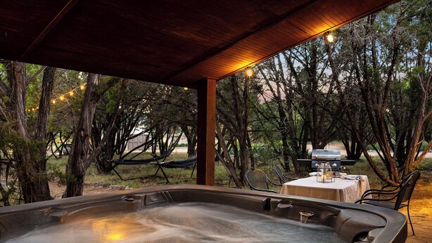 A hot tub under a wooden pergola with string lights, surrounded by trees and a barbecue setup. - Summer Riverfront | 5 Acres | Hot Tub | Sleeps 17 (Wimberley)