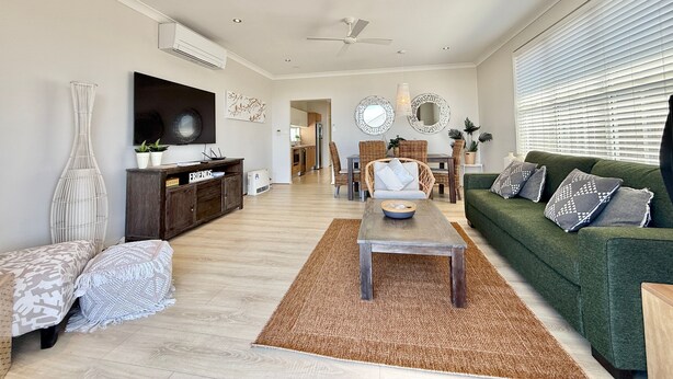 Una sala de estar con un sofá, una mesa de centro y un televisor. Hay un comedor con sillas y una mesa. La habitación tiene un ventilador de techo y una ventana con persianas.