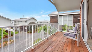 Una terraza de madera con barandilla blanca, un espacio de descanso con cojines y vistas a casas vecinas.