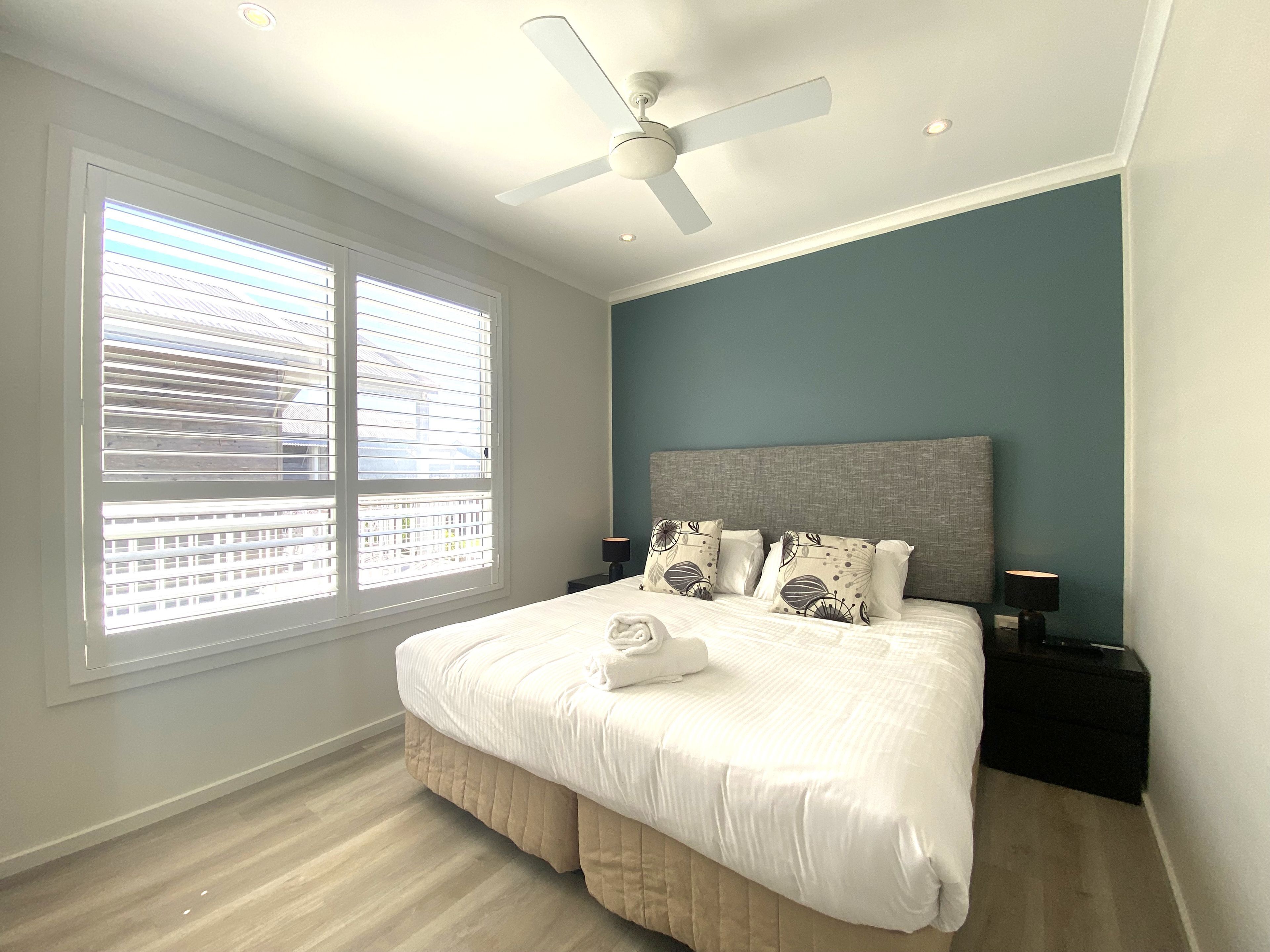 Une chambre à coucher avec un lit, un ventilateur de plafond, une table de chevet et une fenêtre avec des stores.