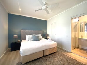 A bedroom with a bed, bedside tables, a ceiling fan, and a bathroom with a mirror and sink. - Azure Tides at Caves Beach Villas (Caves Beach)