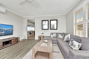 A modern living room with a flat-screen TV, a wooden coffee table, a grey sofa with patterned cushions, and a view of the sea through the window. - Azure Tides at Caves Beach Villas (Caves Beach)
