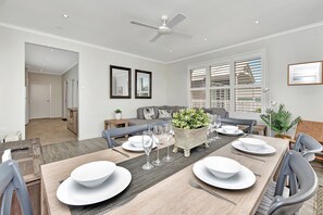 Un coin repas avec une grande table dressée pour six personnes, entourée de chaises. Un ventilateur de plafond est installé au-dessus, et une grande fenêtre avec des stores se trouve sur le côté.