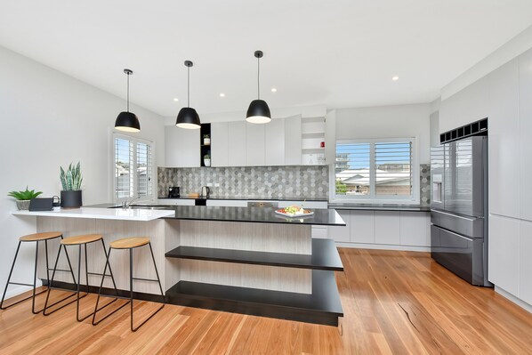 A modern kitchen with a large island, bar stools, pendant lights, and a stainless steel refrigerator.