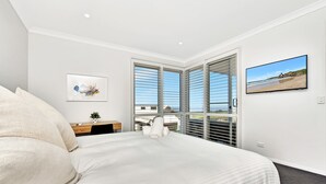 A bedroom with a bed, a desk, a chair, a potted plant, and a window with blinds. - Bombora Beach House at Caves Beach (Caves Beach)