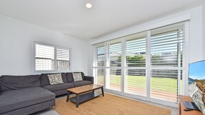 A modern living room with a grey sectional sofa, a wooden coffee table, and a large window with white blinds. - Bombora Beach House at Caves Beach (Caves Beach)