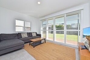 A modern living room with a grey sectional sofa, a wooden coffee table, and a large window with white blinds. - Bombora Beach House at Caves Beach (Caves Beach)