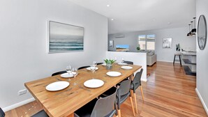 A modern dining area with a wooden table set for four, a large painting of the ocean, and a view into the kitchen area. - Bombora Beach House at Caves Beach (Caves Beach)