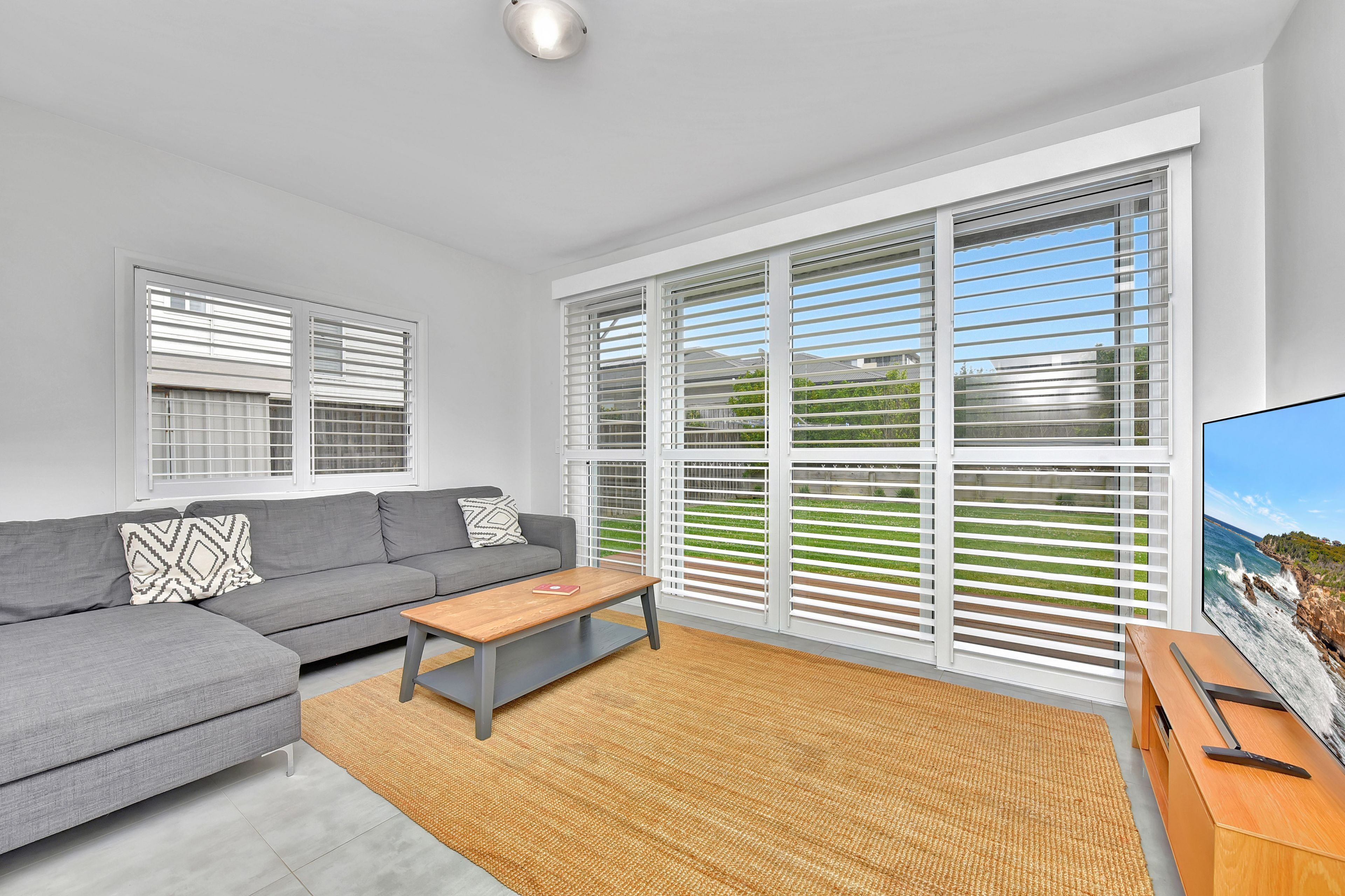 A modern living room with a grey sofa, a wooden coffee table, and a large window with white blinds.