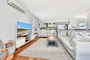A modern living room with a flat-screen TV, a sofa, a coffee table, and a dining area with pendant lights.