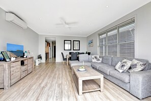 A modern living room with a grey sofa, wooden coffee table, and a dining area with chairs. - Mariner's Rest at Caves Beach Villas (Caves Beach)