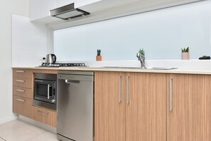 A modern kitchen with a dishwasher, oven, and stove, wooden cabinets, and a white countertop. - Mariner's Rest at Caves Beach Villas (Caves Beach)