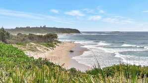 A coastal landscape with a sandy beach, ocean waves, and a grassy hill with trees. - Mariner's Rest at Caves Beach Villas (Caves Beach)