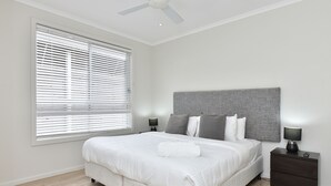 A bedroom with a bed, nightstands, a ceiling fan, and a window with blinds. - Mariner's Rest at Caves Beach Villas (Caves Beach)