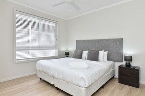 A bedroom with a bed, nightstands, a ceiling fan, and a window with blinds. - Mariner's Rest at Caves Beach Villas (Caves Beach)