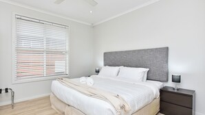 Een moderne slaapkamer met een groot bed, een grijs hoofdeinde, witte lakens en een nachtkastje met een lamp.