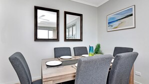 A dining area with a wooden table, six grey upholstered chairs, two wall mirrors, and a framed beach painting. - Mariner's Rest at Caves Beach Villas (Caves Beach)