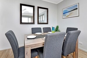 A dining area with a wooden table, six grey upholstered chairs, two wall mirrors, and a framed beach painting. - Mariner's Rest at Caves Beach Villas (Caves Beach)