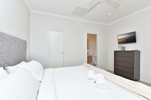 A modern bedroom with a bed, a dresser, a television, and a bathroom visible through an open door. - Mariner's Rest at Caves Beach Villas (Caves Beach)