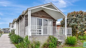 Een gelijkvloers houten huis met een balkon, omringd door groen en een geplaveid pad.