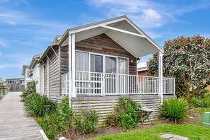 Une maison en bois d’un seul étage, dotée d’un balcon, entourée de verdure et bordée d’un chemin pavé.