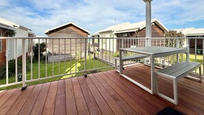 Une terrasse en bois, agrémentée d’une table et de chaises, entourée d’une balustrade blanche, et d’où l’on peut admirer une cour verdoyante avec des maisons en arrière-plan.