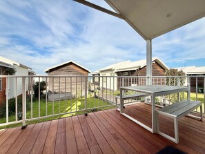 Une terrasse en bois, agrémentée d’une table et de chaises, entourée d’une balustrade blanche, et d’où l’on peut admirer une cour verdoyante avec des maisons en arrière-plan.