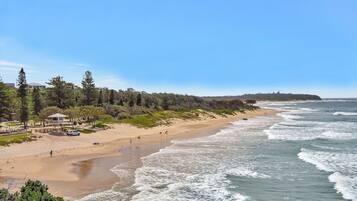 A beach with a sandy shore, waves, and a forested area with buildings in the background.