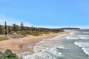 Una playa con arena, olas y una zona boscosa con edificios al fondo.