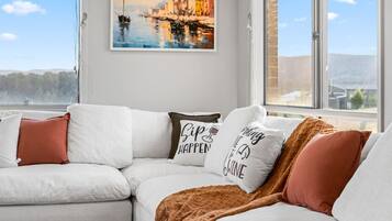 Un salon moderne avec un canapé blanc, un tableau représentant un paysage côtier et une fenêtre donnant sur l’extérieur.