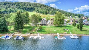 En ejendom ved søen med fortøjede både, en græsplæne og huse med en skovklædt bakke i baggrunden.