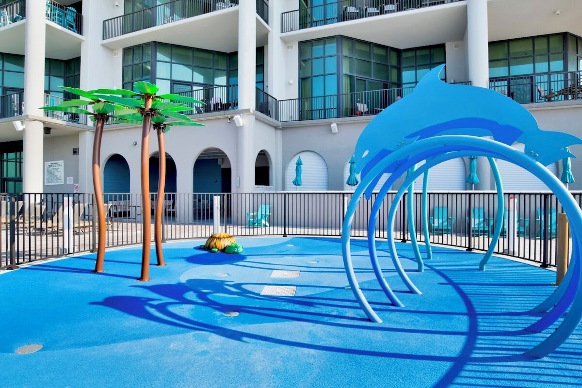 Zona de juegos infantiles con piso azul, una gran escultura de delfín azul y dos palmeras.