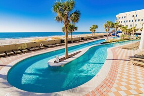 A swimming pool with palm trees, a beach, and a hotel building. - The Oasis 2005 | Beach front! Spacious, clean, slide, lazy river, pickle ball, bar/grill! (Orange Beach)