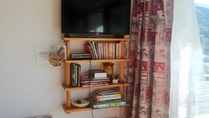 A room with a mounted TV, bookshelves, a radiator, and patterned curtains. - Studio 'Pathabac' with Mountain View and Balcony (Auris)