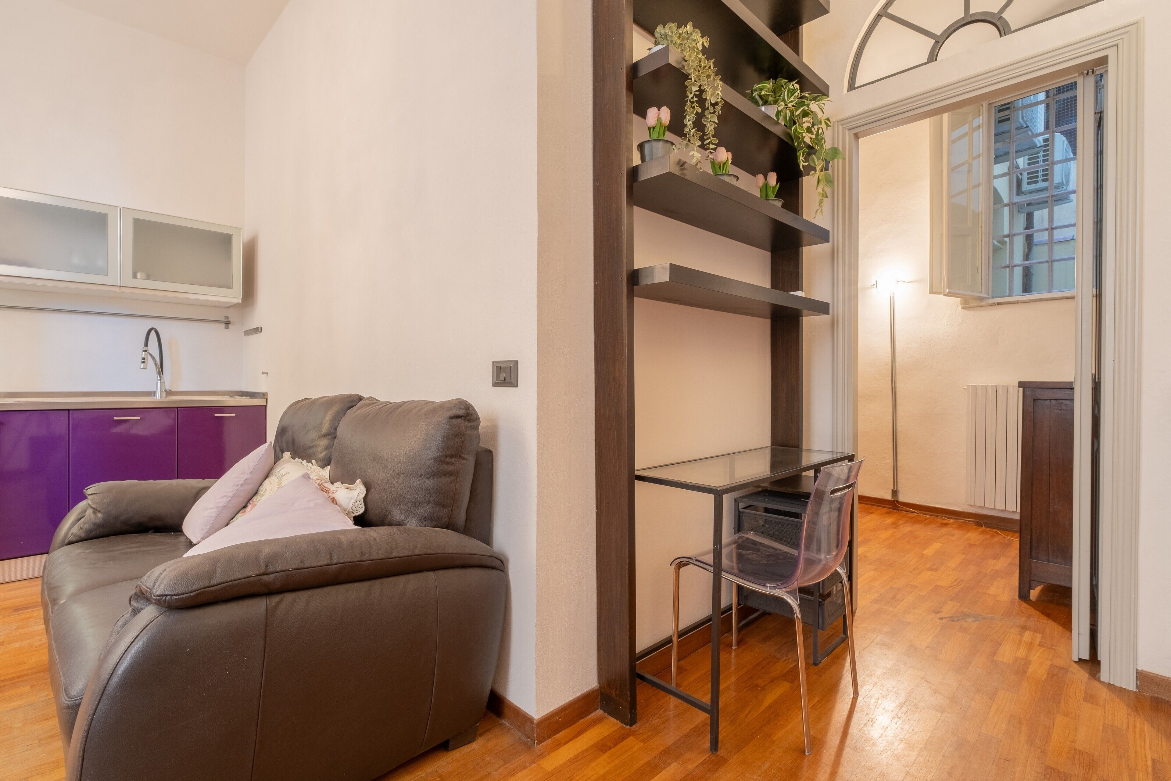 Una sala de estar moderna con un sofá, una mesa de comedor con sillas, una zona de cocina con armarios morados y una ventana con vistas.