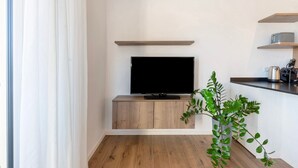 Ett modernt vardagsrum med en platt-TV placerad på ett träskåp, en krukväxt och inbyggda hyllor med köksredskap.