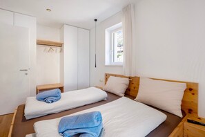 A bedroom with two beds, a wooden headboard, a window with curtains, a bedside table, and a hanging clothes rack.